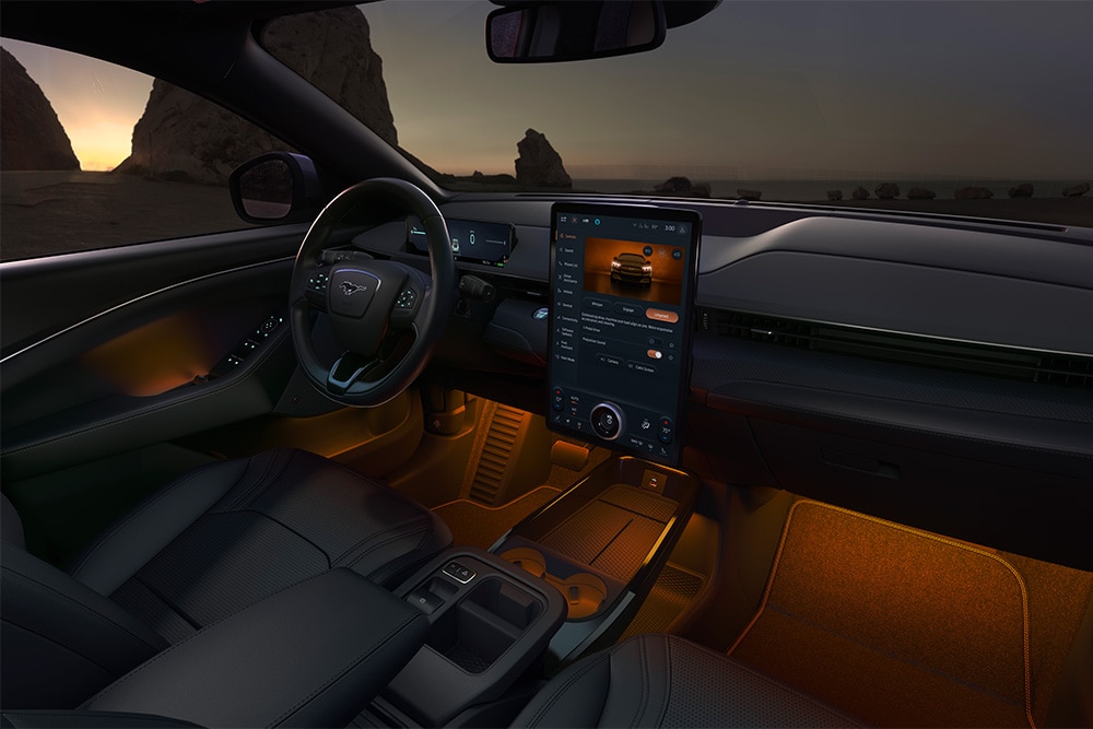 Ford Mustang Mach-E Innenraum. Ansicht mit LED-Ambientebeleuchtung in Orange und Blick auf das Lenkrad und den Ford SYNC 4-Touchscreen.