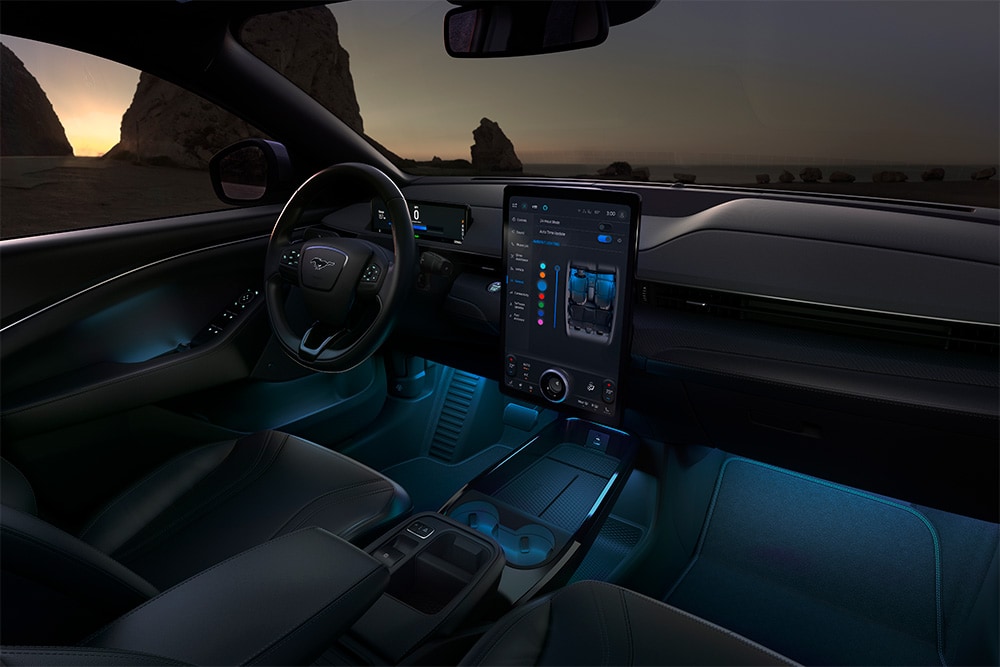 Ford Mustang Mach-E Innenraum. Ansicht mit LED-Ambientebeleuchtung in Blau und Blick auf das Lenkrad und den Ford SYNC 4-Touchscreen.