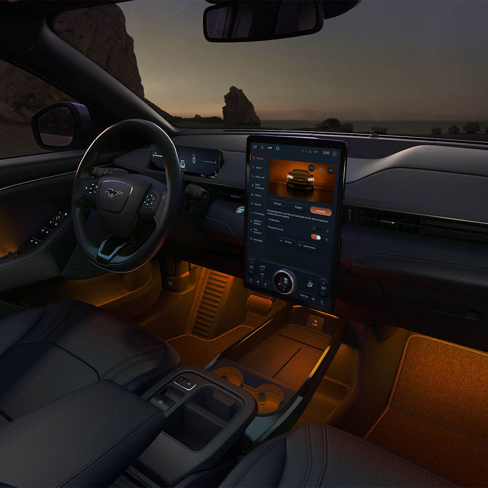 Ford Mustang Mach-E Innenraum. Ansicht mit LED-Ambientebeleuchtung in Orange und Blick auf das Lenkrad und den Ford SYNC 4-Touchscreen.