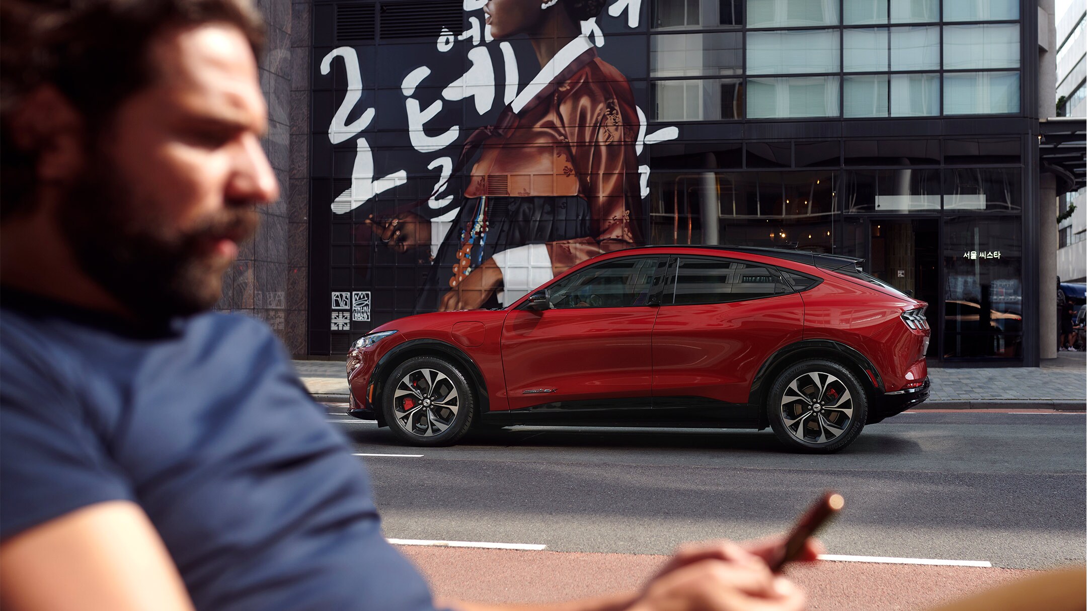 Ford Mustang Mach-E in Rot. Seitenansicht, parkend vor einem modernen Gebäude. Im Vordergrund ein Mann der auf sein Smartphone schaut