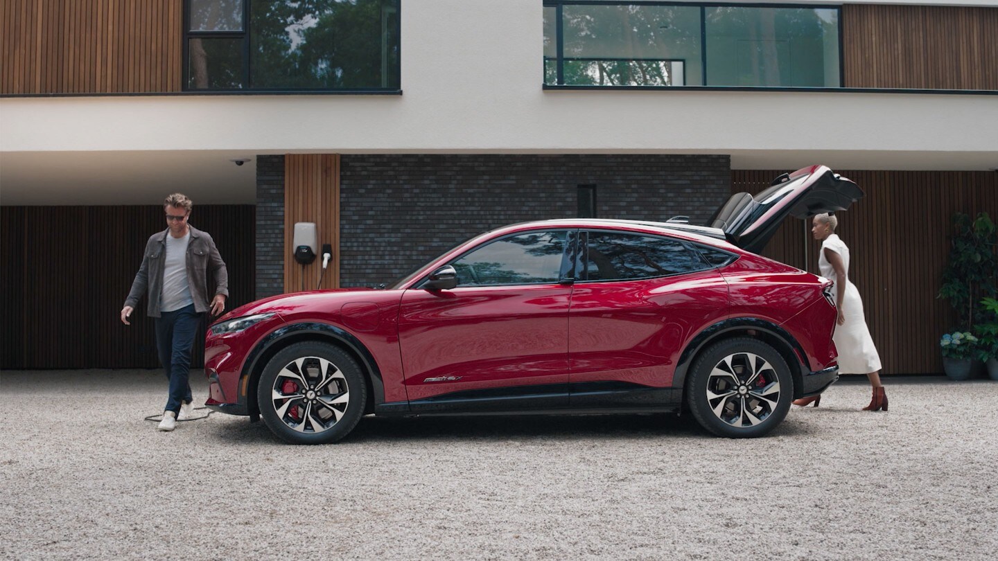 Ford Mustang Mach-E AWD in Rot. Seitenansicht, parkend vor einem modernen Gebäude mit geöffneter Heckklappe