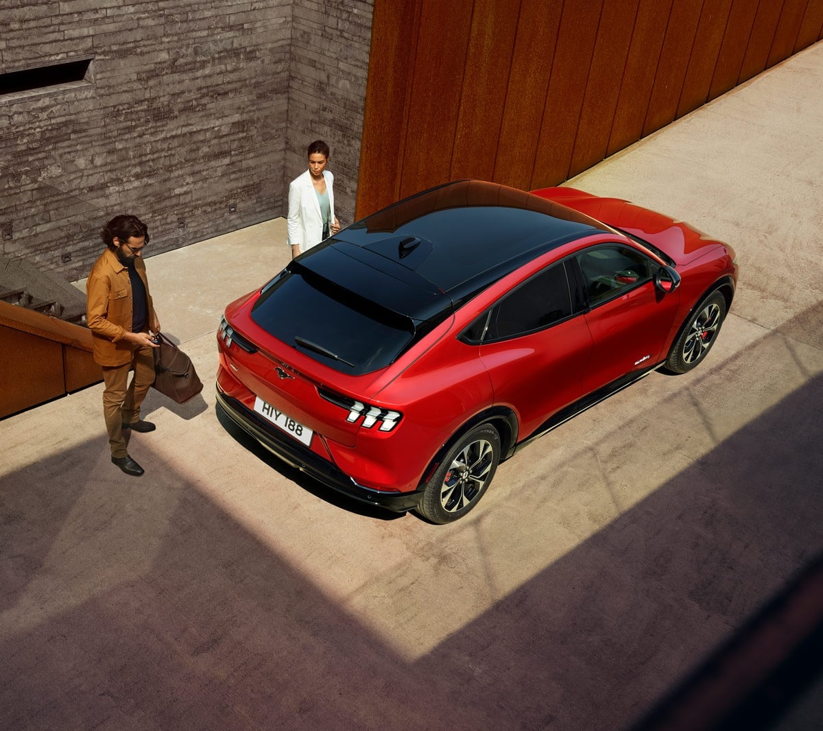 Ford Mustang Mach-E in Rot. Dreiviertelansicht von oben, parkend vor einem modernen Gebäude. Frau und Mann stehen neben dem Fahrzeug