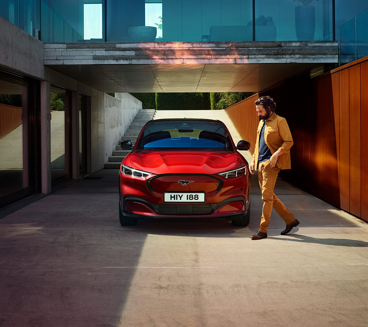 Ford Mustang Mach-E in Rot. Frontansicht, parkend vor einem modernen Gebäude. Mann geht um das Fahrzeug herum