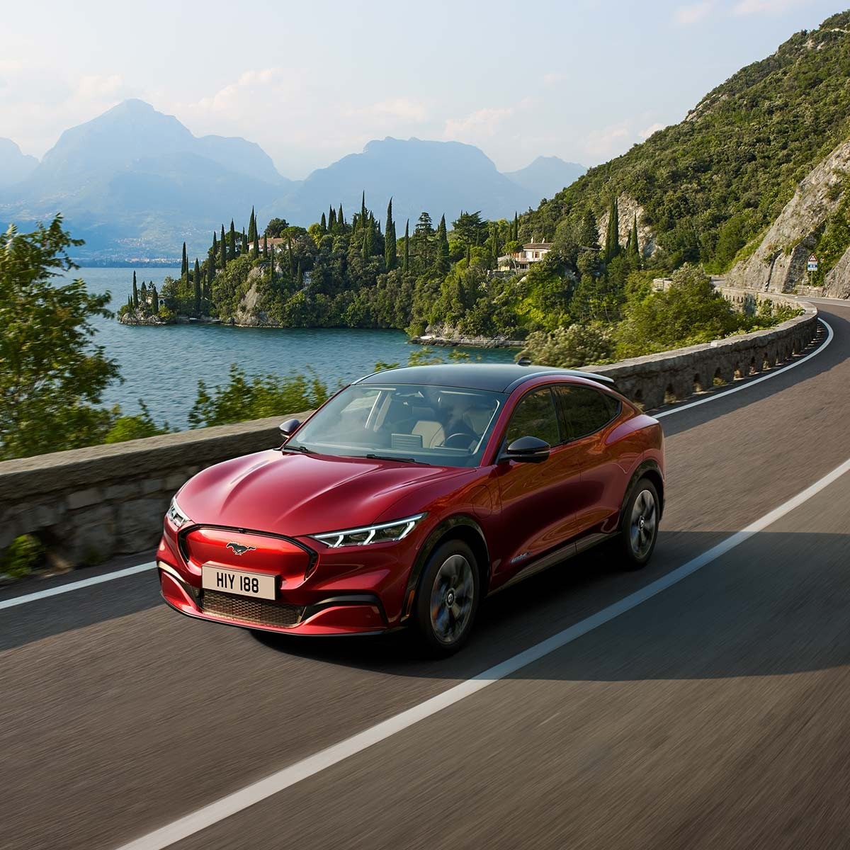 Ford Mustang Mach-E in Rot. Dreiviertelansicht von vorne, fahrend auf einer Straße am Ufer eines Sees