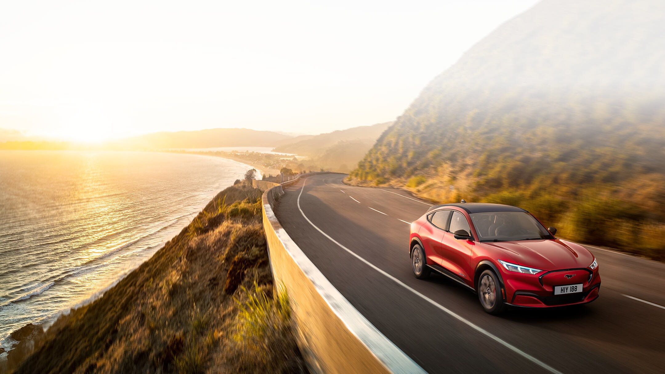Ford vollelektrische Fahrzeuge. Ford Mustang Mach-E in Rot. Seitenansicht, fahrend auf Straße vor See mit baumbewachsenen Ufern