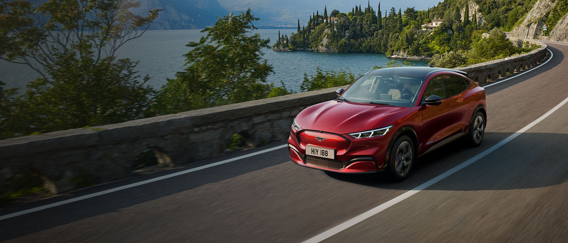 Ford vollelektrische Fahrzeuge. Ford Mustang Mach-E in Rot, Dreiviertelansicht, fahrend auf Bergstraße an See