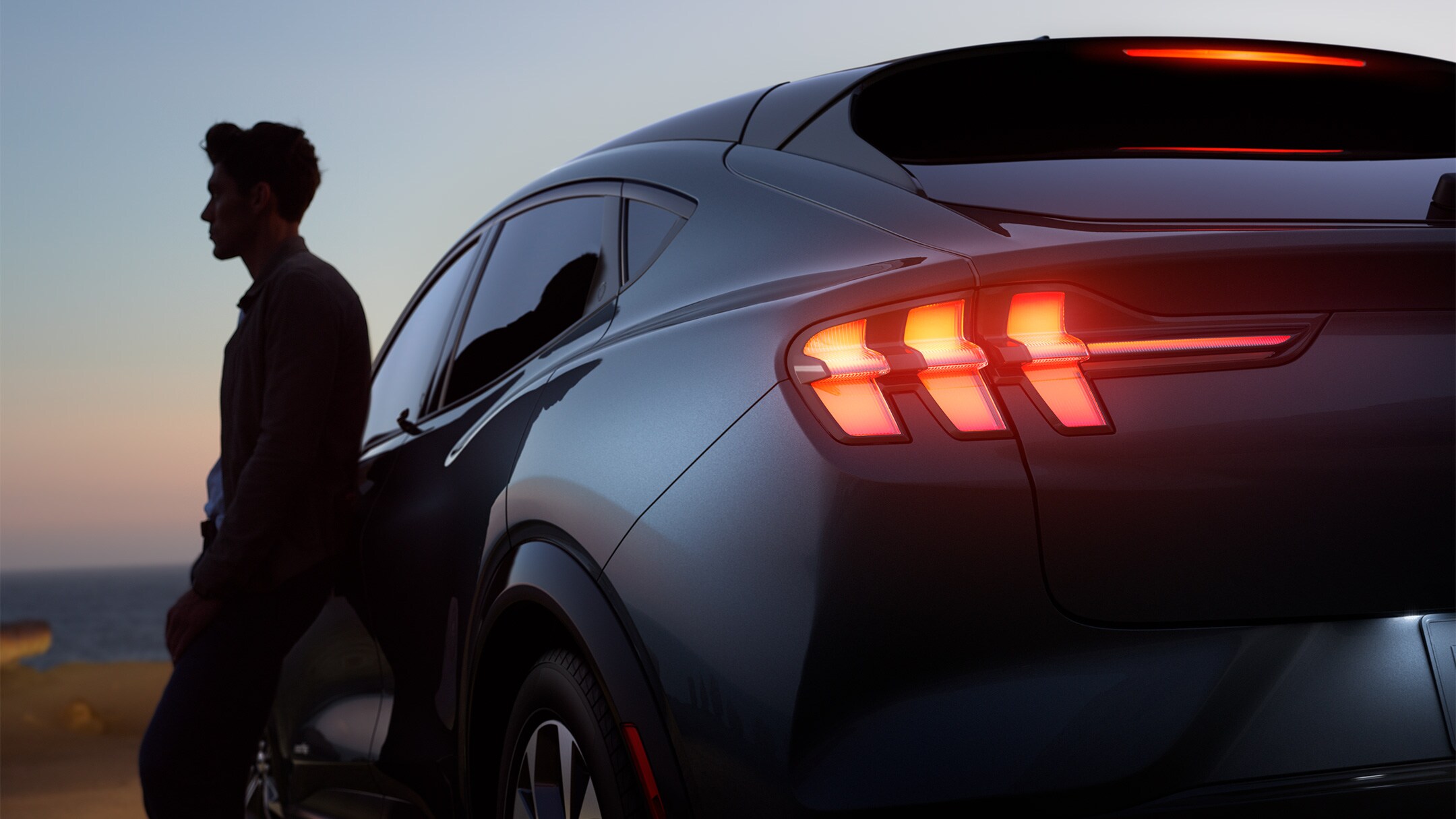 Ford Mustang Mach-E in Grau. Dreiviertelansicht von hinten, parkend bei Abenddämmerung. Mann lehnt am Fahrzeug