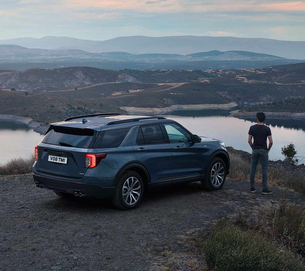 Ford Explorer Plug-in-Hybrid in Blau. Dreiviertelansicht von hinten, parkend in einer hügeligen Landschaft. Mann steht daneben und betrachtet die Landschaft