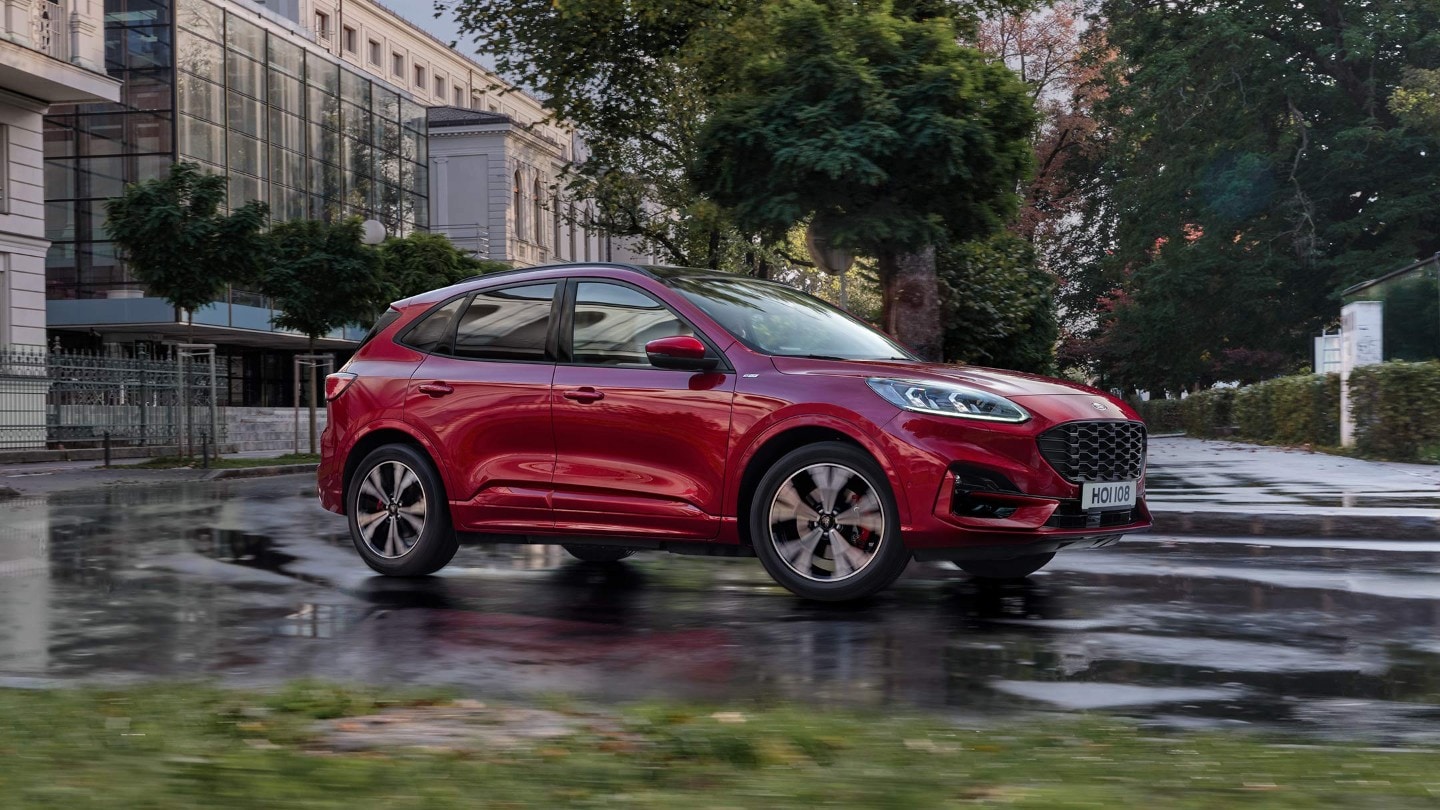 Ford Kuga in Rot. Dreiviertelansicht, durch eine Stadt fahrend von modernen Gebäuden und Bäumen