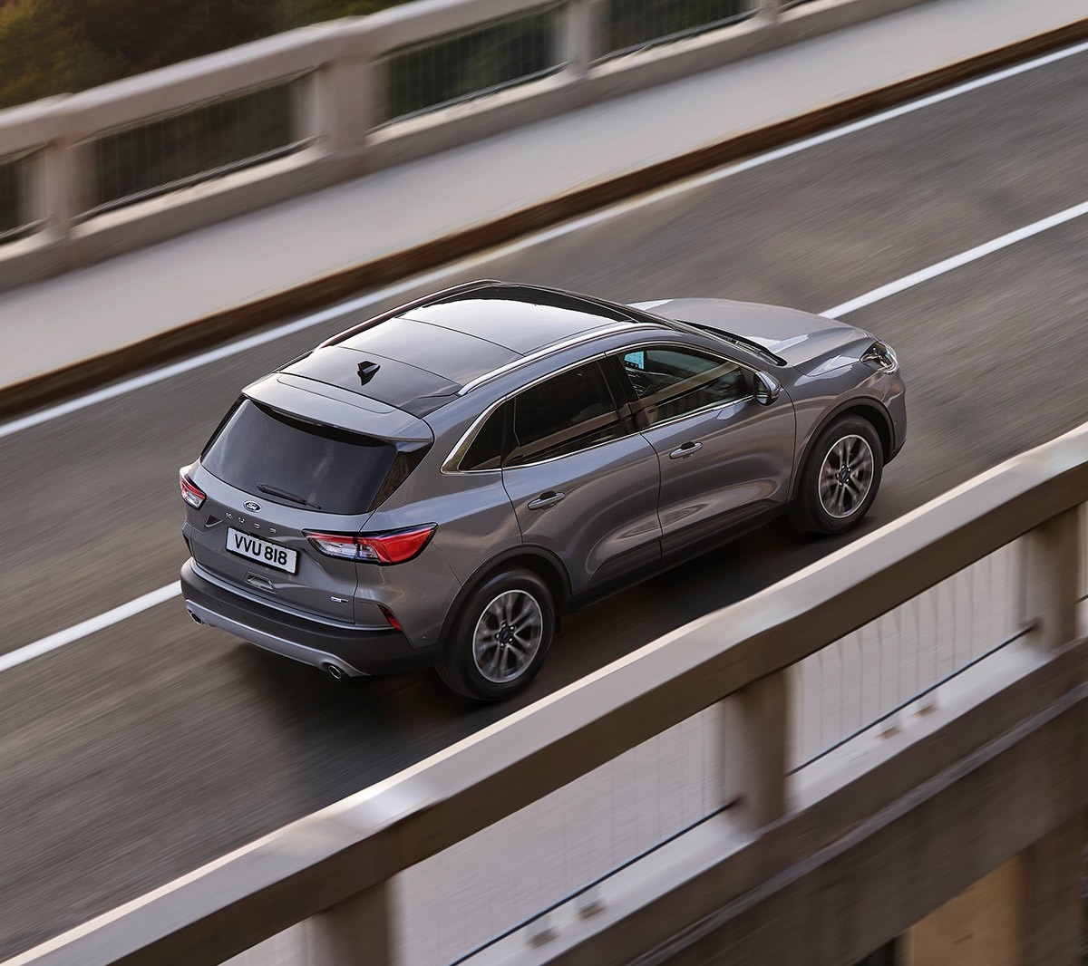 Ford Kuga in Grau. Dreiviertelansicht von oben, fahrend über eine Brücke mit Wald im Hintergrund