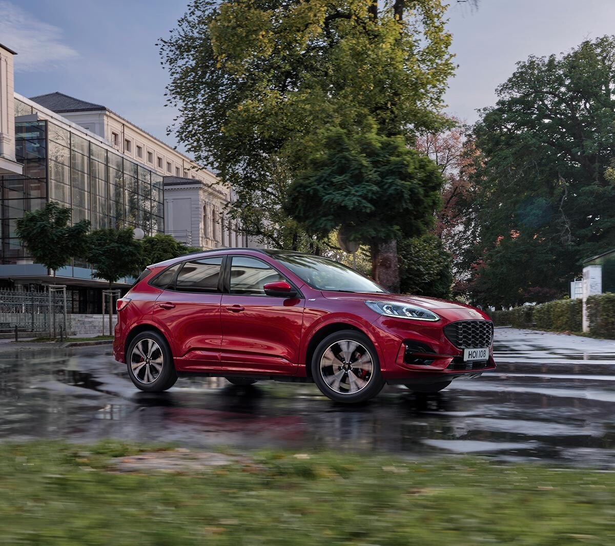 Ford Kuga in Rot. Dreiviertelansicht, fahrend durch eine Stadt vor modernem Gebäude