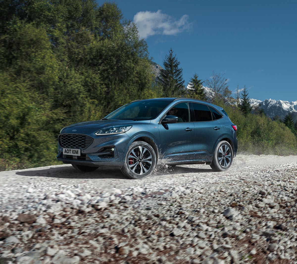 Ford Kuga in Blau. Dreiviertelansicht, fahrend auf einem Schotterweg vor einem Wald