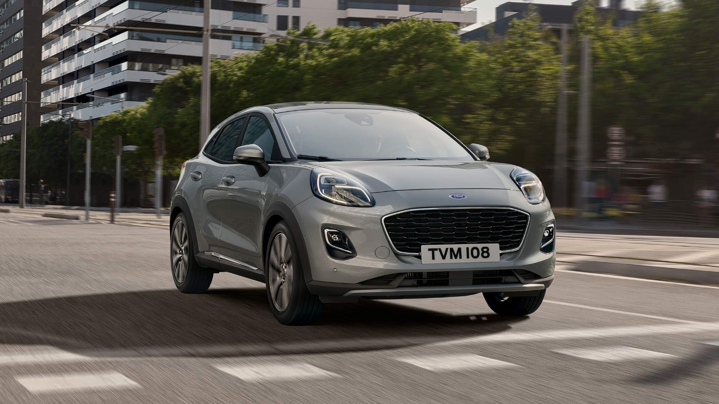 Ford Puma in Grau. Dreiviertelansicht, fahrend auf Straße mit Bäumen und Hochhaus im Hintergrund