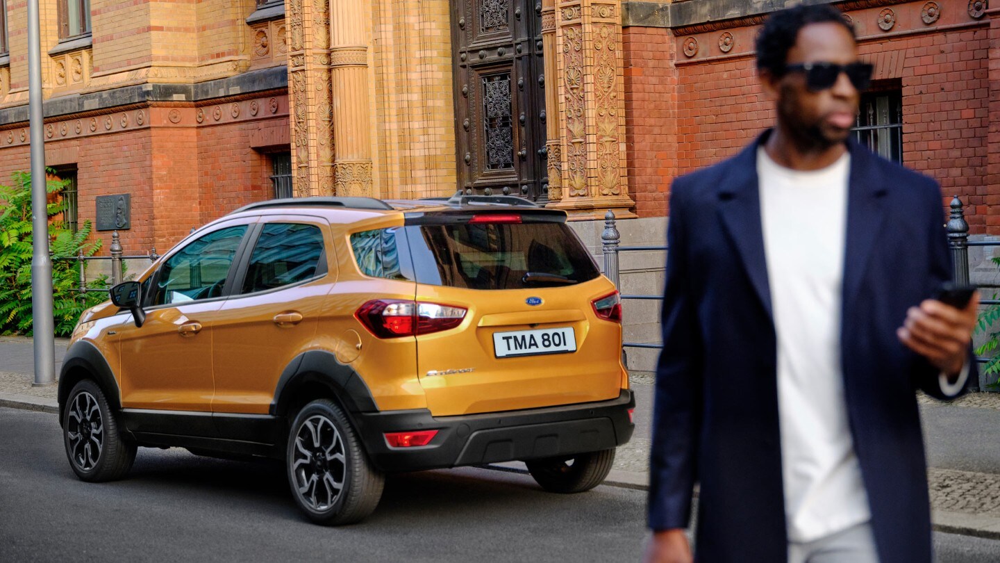 Ford EcoSport Active in Orange. Dreiviertelansicht von hinten, parkend vor einem Gebäude in der Stadt. Mann im Vordergrund schaut auf sein Smartphone