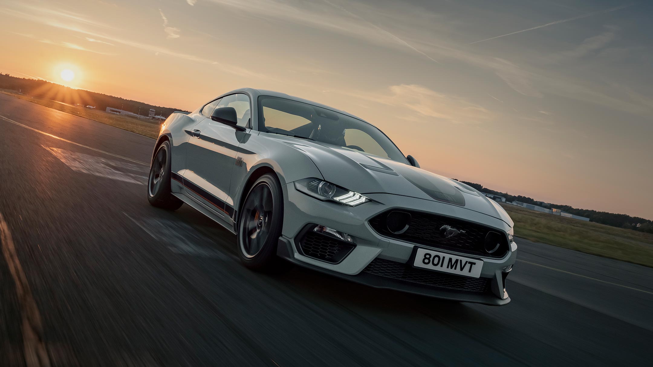 Ford Mustang Mach 1 in Weiß. Dreiviertelansicht von vorne, fahrend auf einer Strecke im Sonnenuntergang
