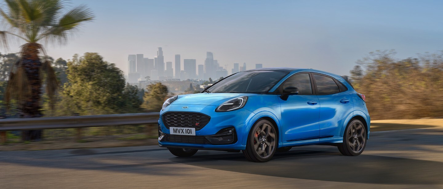 Ford Puma ST in Blau. Dreiviertelansicht, fahrend auf der Straße mit einer Skyline im Hintergrund.