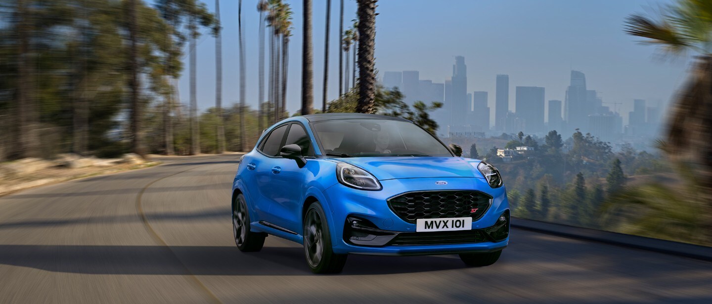 Ford Puma ST in Blau. Frontansicht, fahrend auf der Straße mit einer Skyline im Hintergrund.