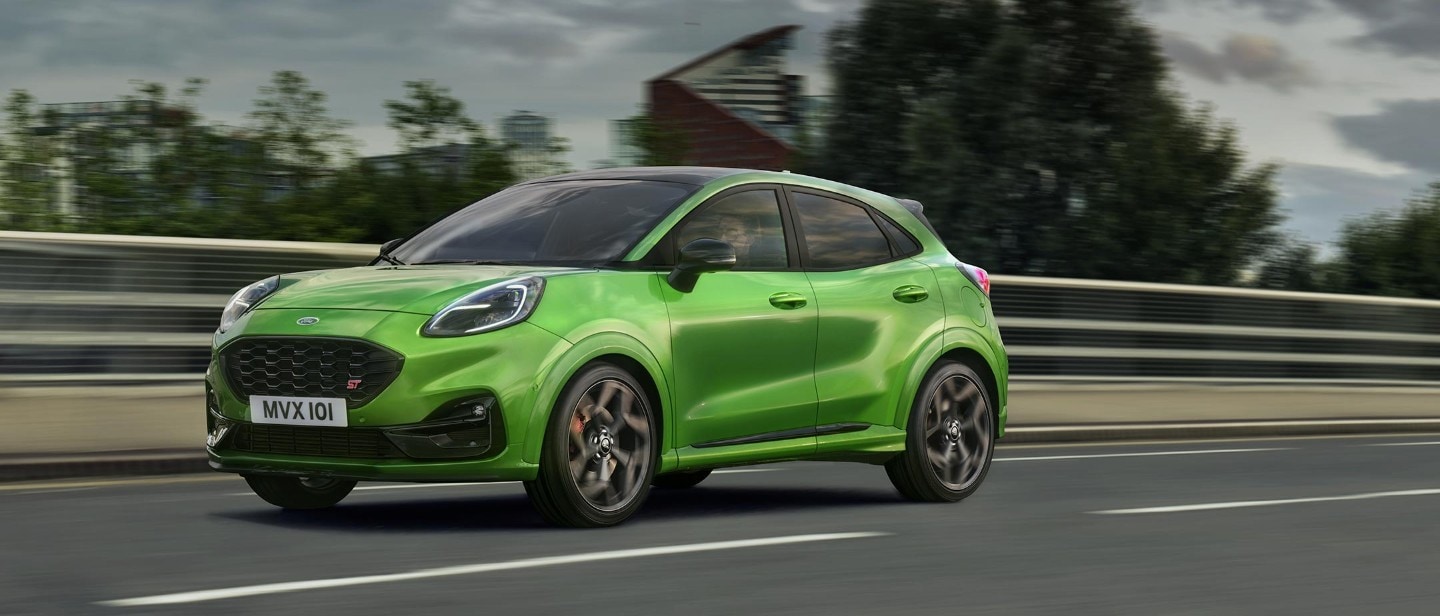 Ford Puma ST in Grün. Dreiviertel-Frontansicht, fahrend auf einer Landstraße mit Hochhäusern im Hintergrund