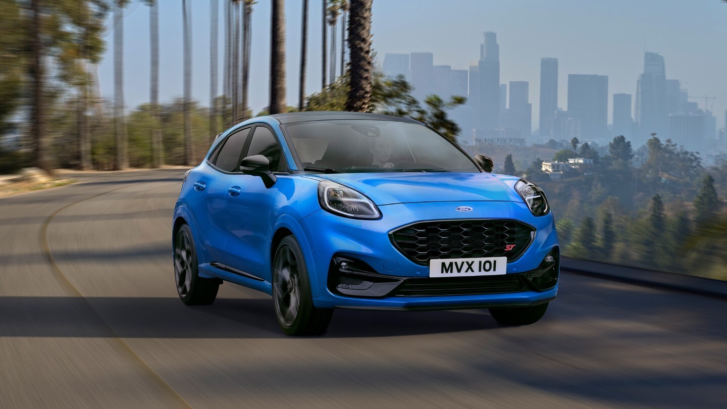 Ford Puma ST in Blau. Frontansicht, fahrend auf der Straße mit einer Skyline im Hintergrund.