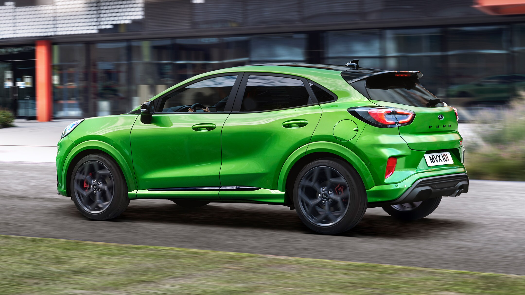 Ford Puma ST in Grün. Dreiviertel-Heckansicht. Auf einer Straße vor modernem Gebäude fahrend