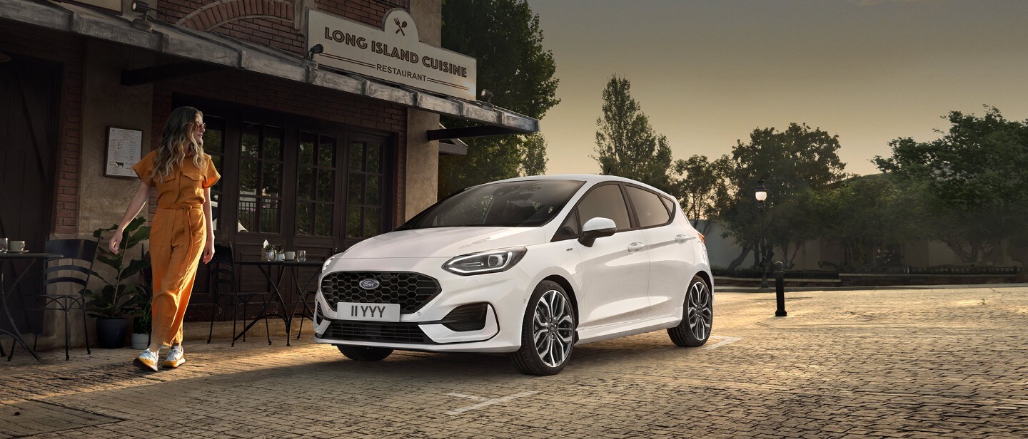 Ford Fiesta in Weiß. Dreiviertelansicht, vor einem Restaurant parkend mit Frau im Vordergrund.