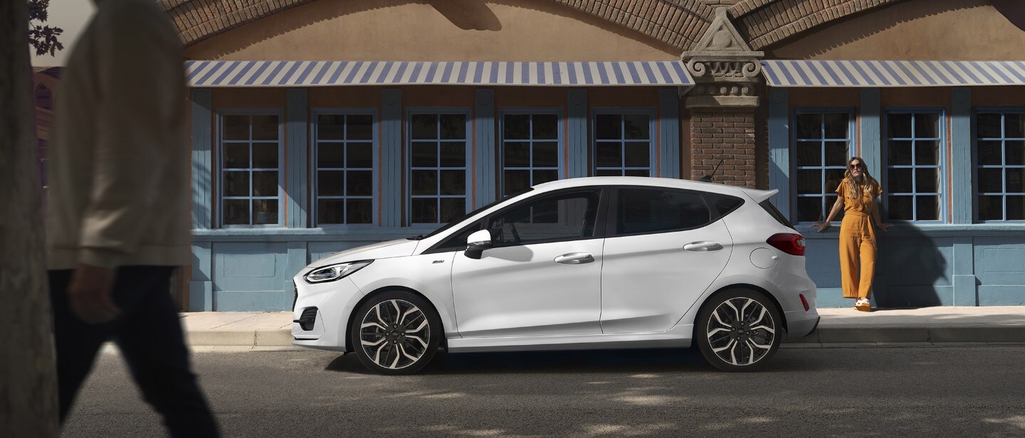Ford Fiesta in Weiß. Dreiviertelansicht, vor einem Restaurant parkend mit Frau im Vordergrund