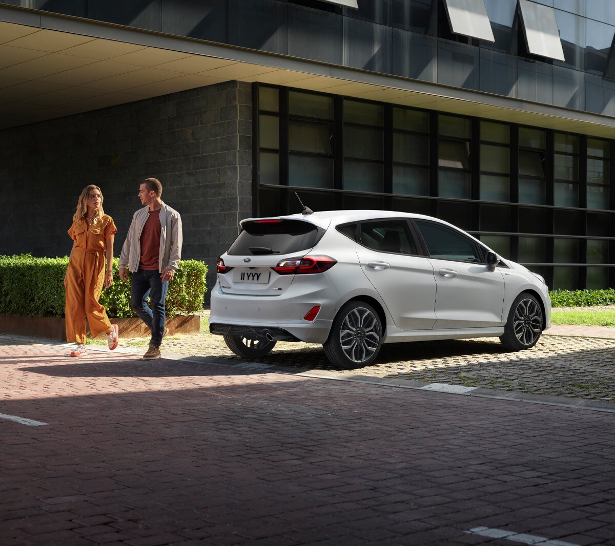Ford Fiesta in Weiß. Heckansicht vor Gebäuden parkend mit zwei Personen daneben.