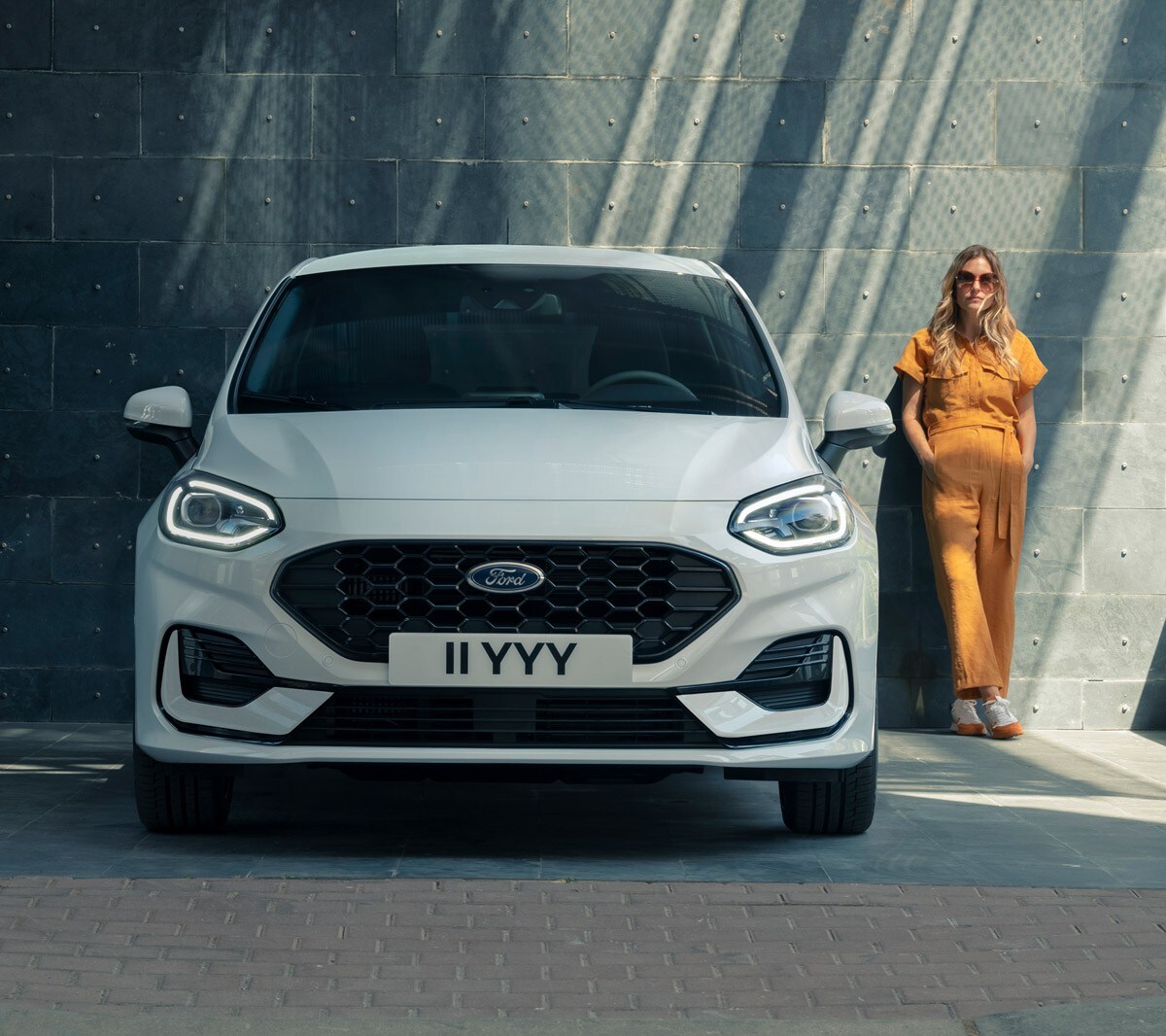 Ford Fiesta in Weiß. Frontansicht, vor einem modernen Gebäude parkend mit Frau im Hintergrund.