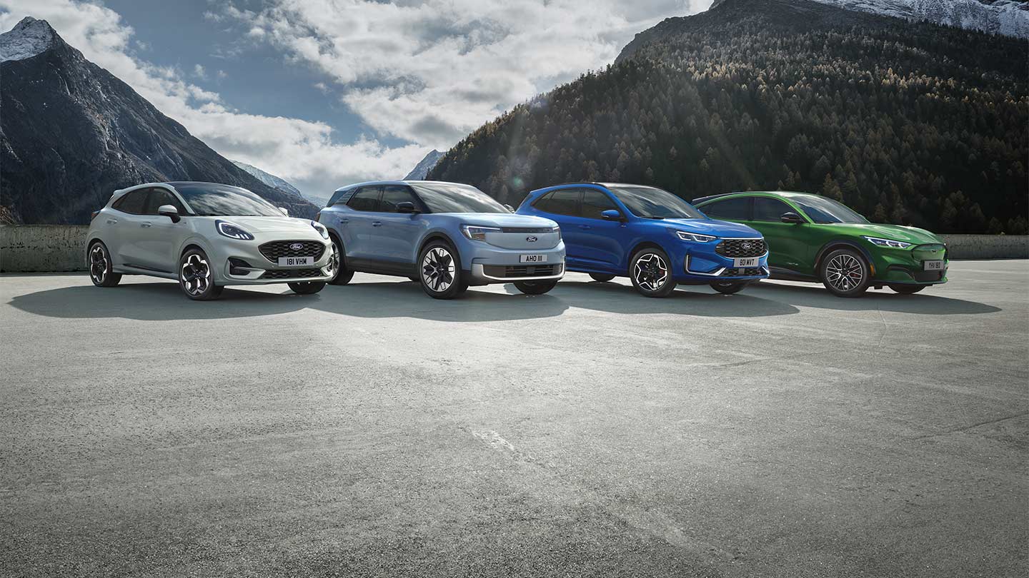 Ford Puma, Ford Explorer, Ford Kuga und Mustang Mach-E stehen nebeneinander auf einem Parkplatz mit Berglandschaft im Hintergrund.