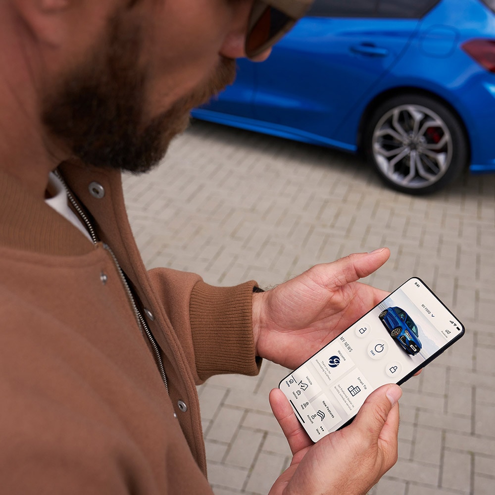 FordPass App. Ein Mann schaut auf sein Smartphone. Im Hintergrund ein blauer Ford Focus.
