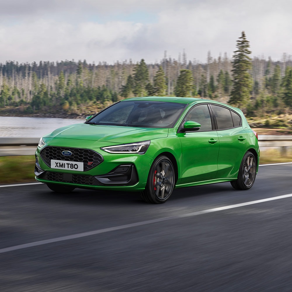 Ford Focus ST in Grün. Dreiviertelansicht, fahrend auf einer Straße. Bäume im Hintergrund.