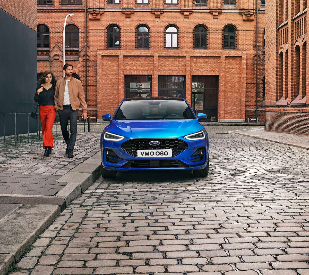 Ford Focus ST-Line in Blau. Frontansicht, in einer industriellen Gegend parkend. Zwei Personen gehen daneben.