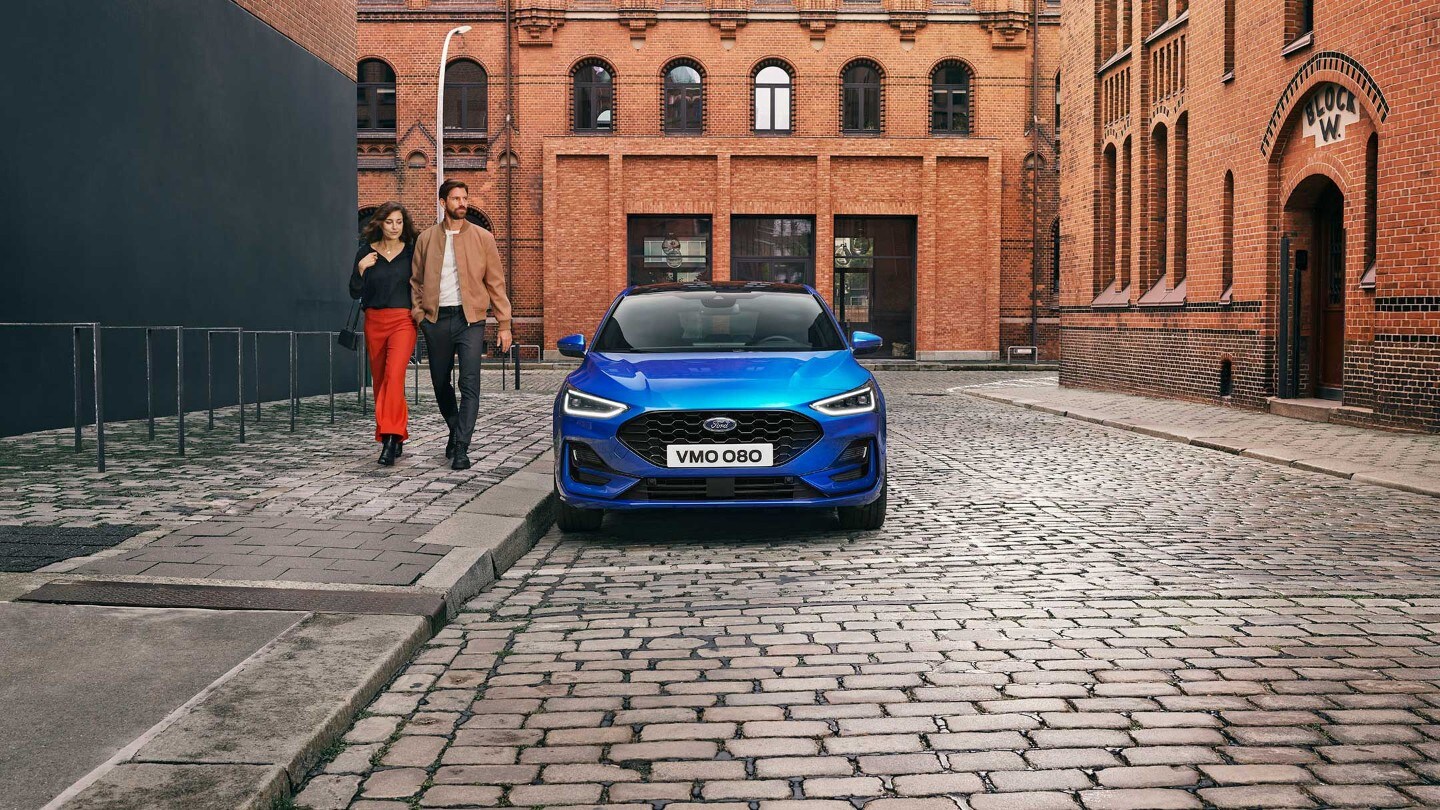 Ford Focus ST-Line in Blau. Frontansicht, in einer industriellen Gegend parkend. Zwei Personen gehen daneben.