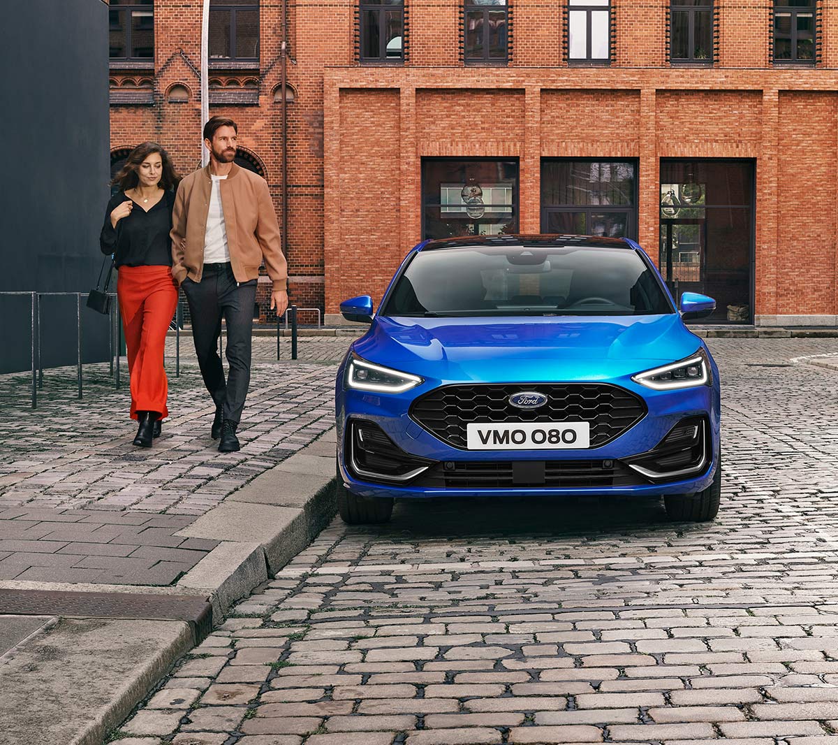 Ford Focus in Blau. Frontansicht, in einer industriellen Gegend parkend. Zwei Personen gehen daneben.