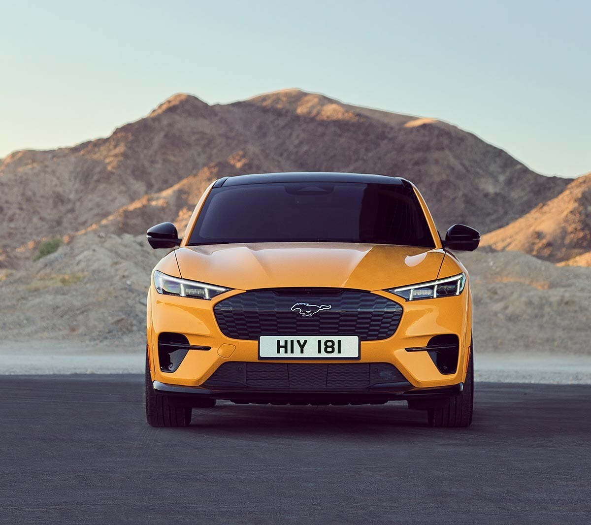Ford Mustang Mach-E GT in Orange. Frontansicht, parkend auf einem Platz mit Bergen im Hintergrund