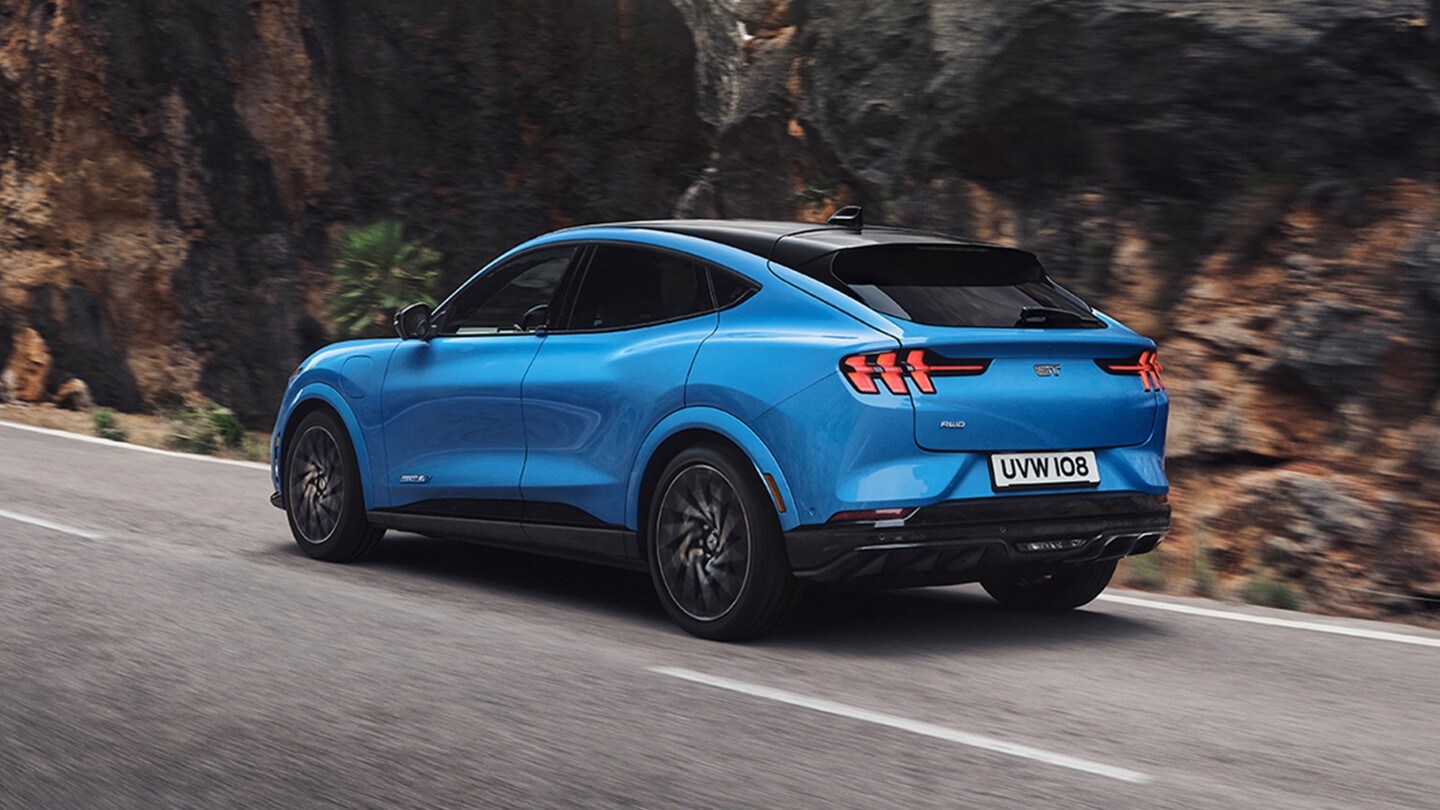 Ford Mustang Mach-E GT in Blau. Heckansicht, auf einer Landstraße fahrend.