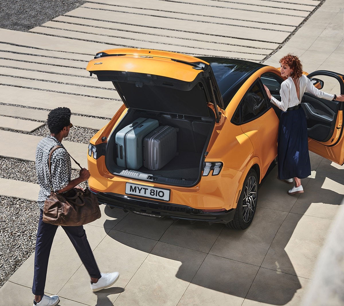 Ford Mustang Mach-E GT in Orange. Heckansicht, zwei Personen nutzen die sensorgesteuerte Heckklappe.
