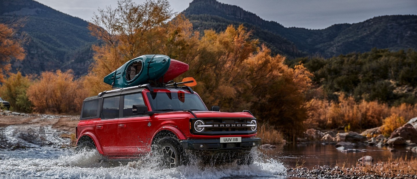 Ford Bronco in Rot. Frontansicht, fahrend in den Bergen durch einen Fluss.