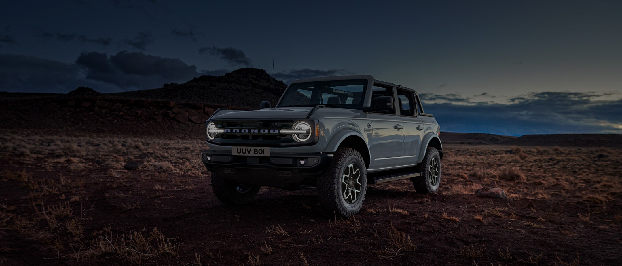 Ford Bronco in Grau. Dreiviertel-Frontansicht, fahrend in der Natur im Sonnenuntergang.