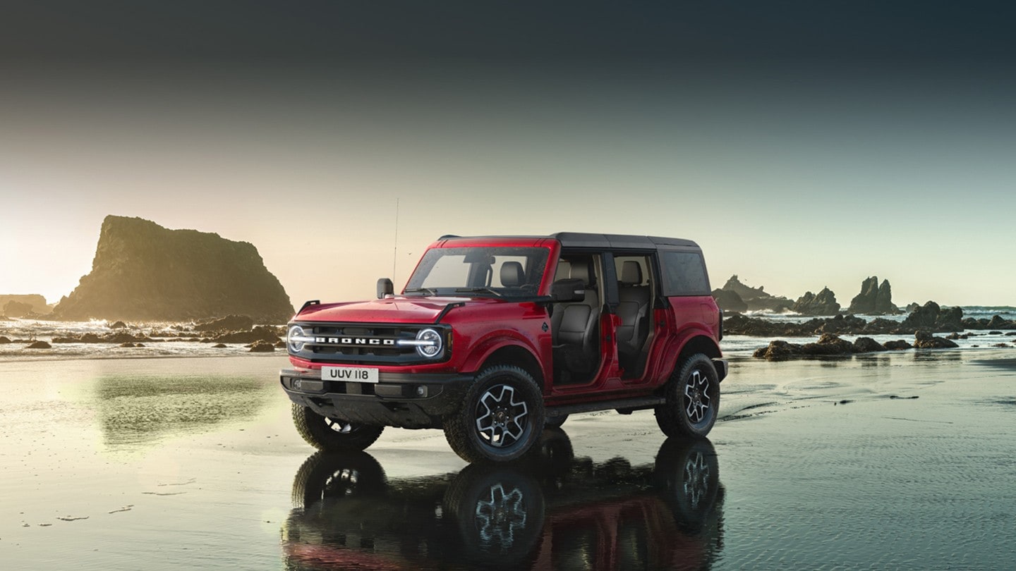 Ford Bronco in Rot. Frontansicht, fahrend in den Bergen durch einen Fluss.