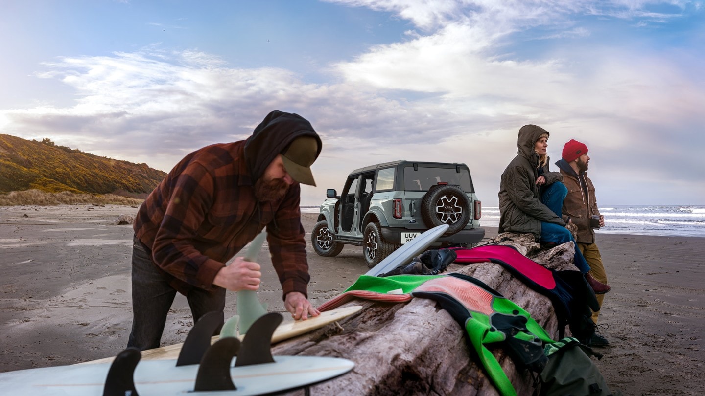 Ford Bronco in Grau. Heckansicht, parkend am Strand mit Surfer im Vordergrund.  
