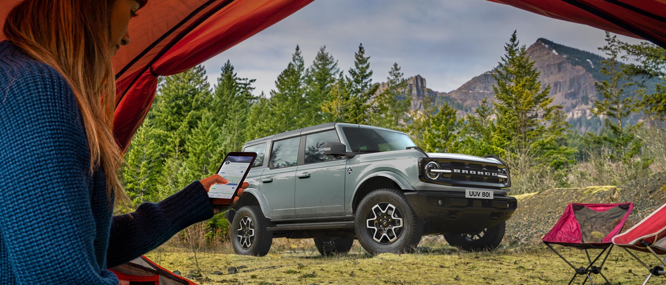 Ford Bronco in Grau. Dreiviertelansicht, stehend vor einem Zelt in dem eine Frau mit Smartphone sitzt