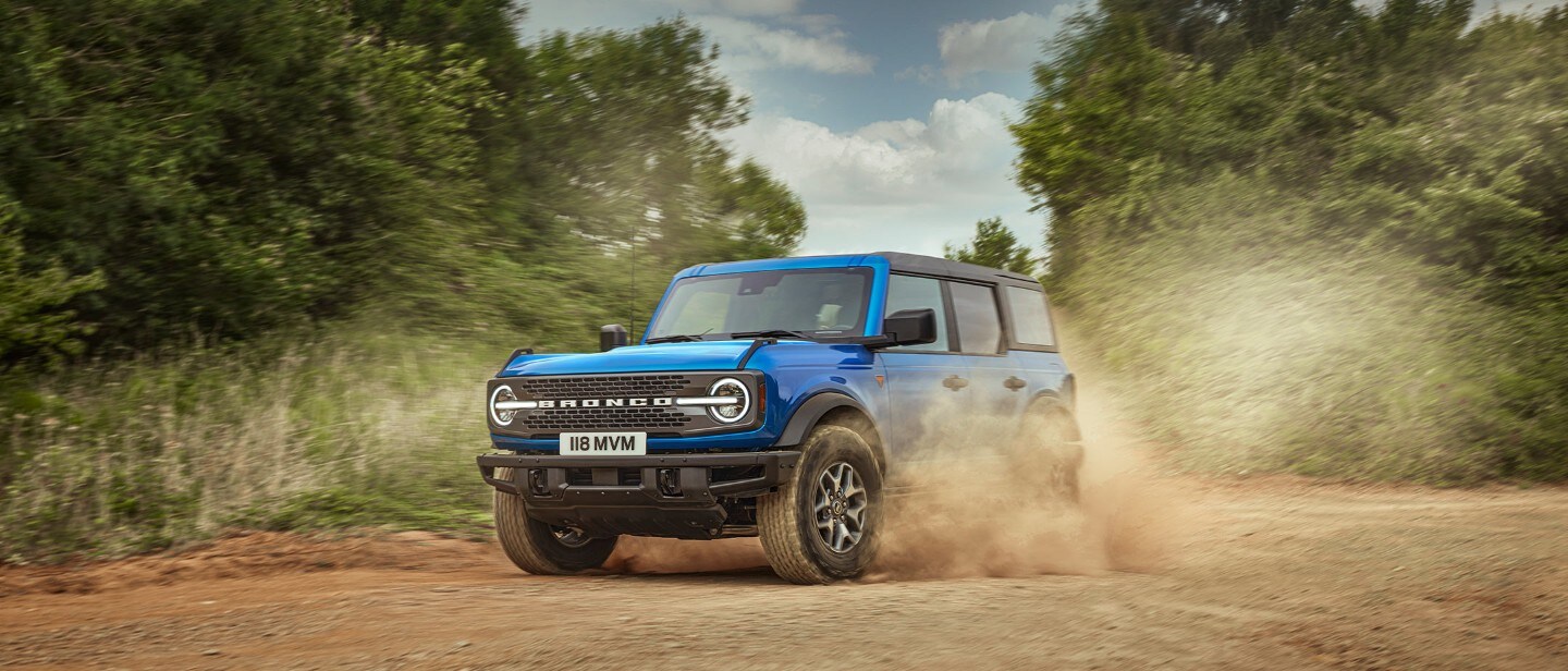 Ford Bronco in Blau. Dreiviertelansicht, fahrend auf staubigem Feldweg