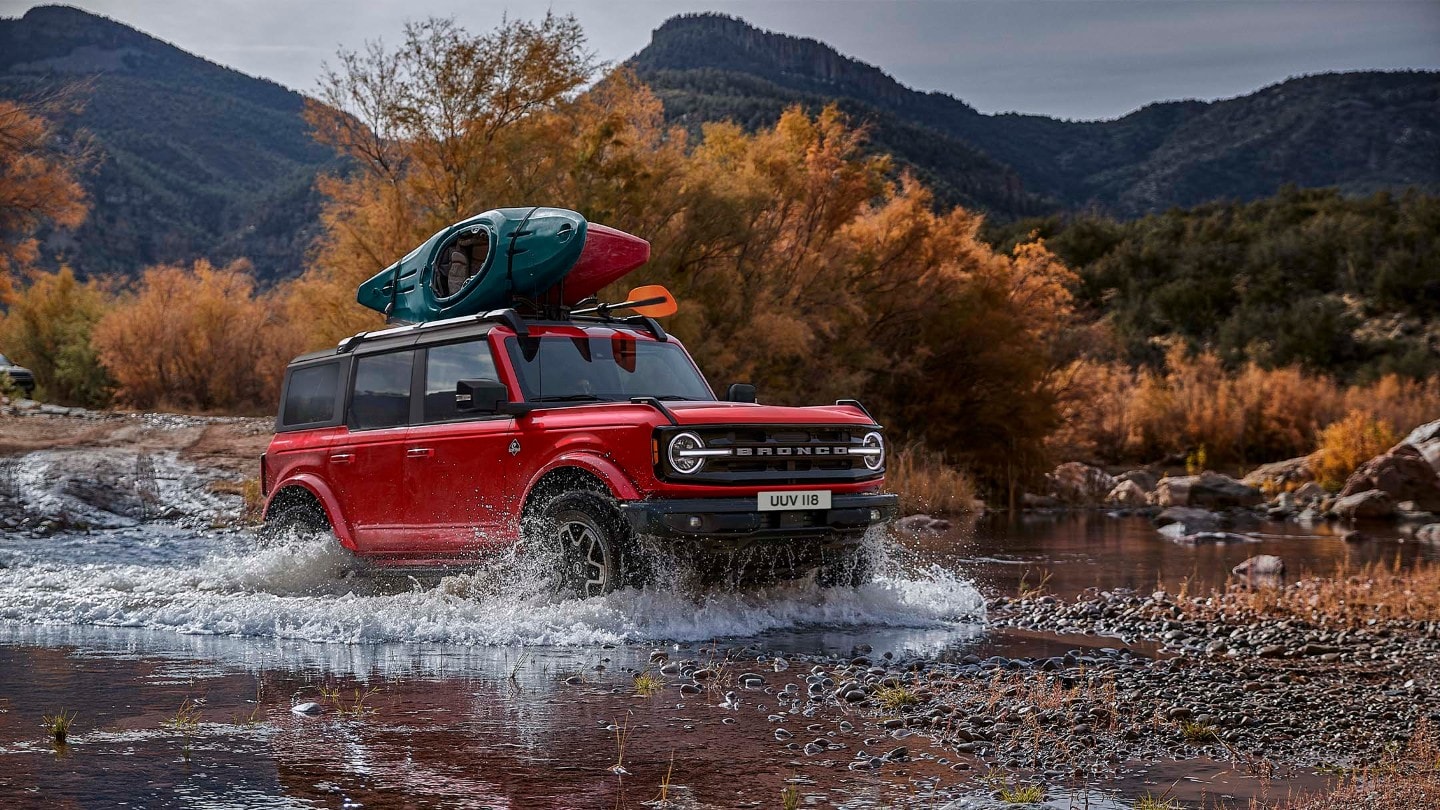 Ford Bronco in Rot. Dreiviertelansicht, fahrend in felsiger Landschaft durch einen Wasserlauf