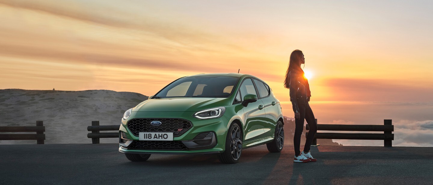 Ford Fiesta ST in Grün. Dreiviertelansicht, Frau betrachtet den Sonnenuntergang.