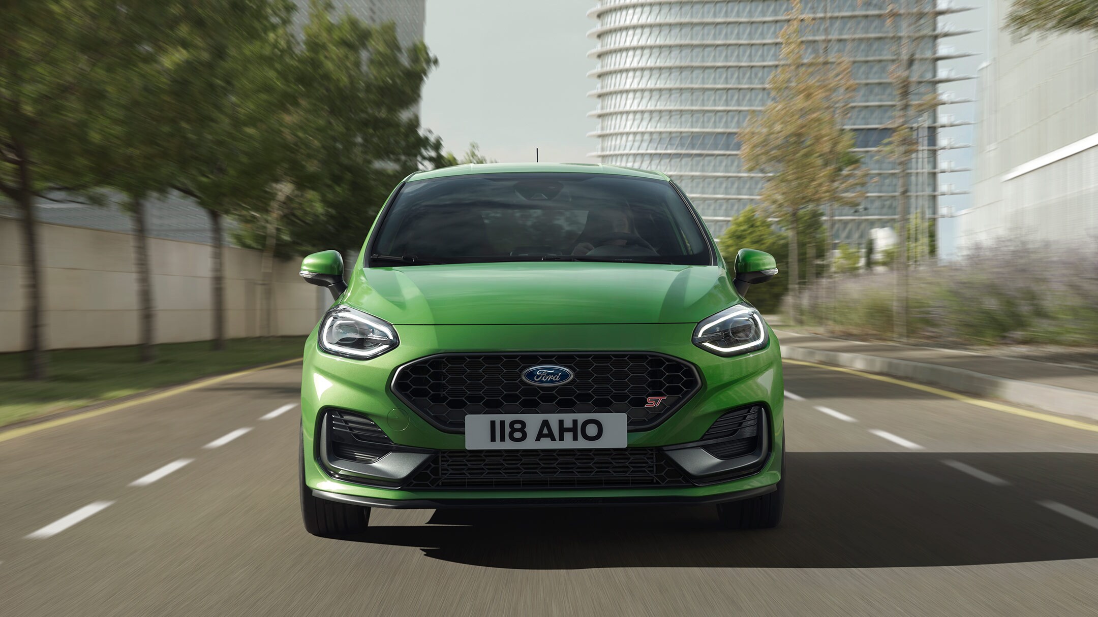 Ford Fiesta ST in Grün. Frontansicht, auf Straße vor Bäumen und modernem Gebäude.