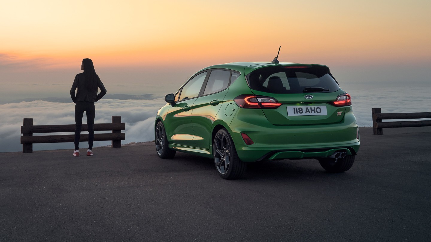 Ford Fiesta ST in Grün. Heckansicht, am Meer parkend mit Frau bei Sonnenuntergang.
