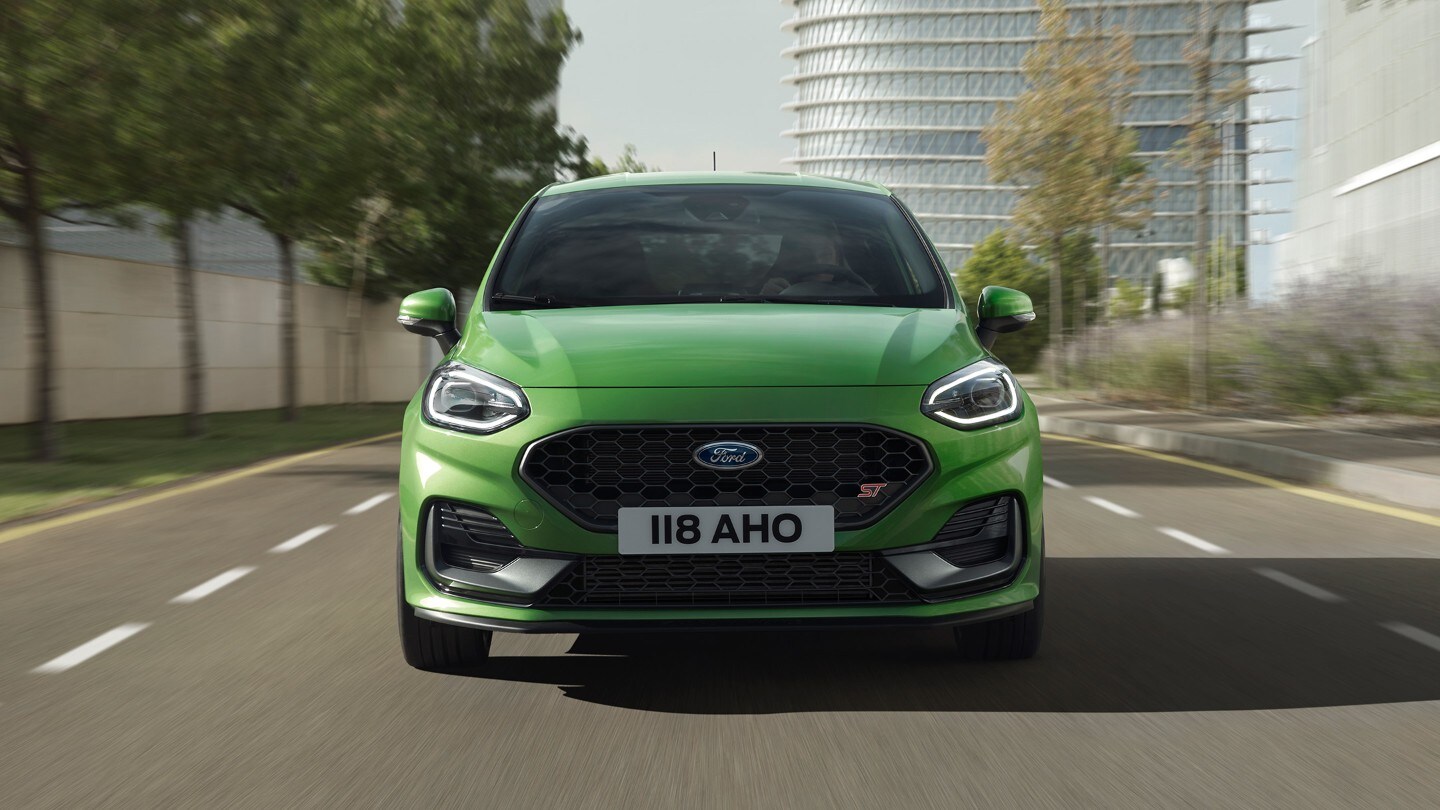 Ford Fiesta ST in Grün. Frontansicht, auf Straße vor Bäumen und modernem Gebäude.