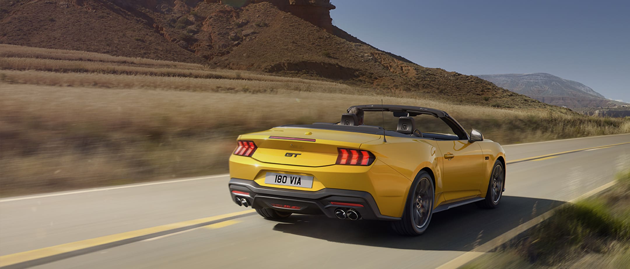 Ford Mustang in Gelb. Heckansicht, fährt auf einer Straße mit Bergen im Hintergrund