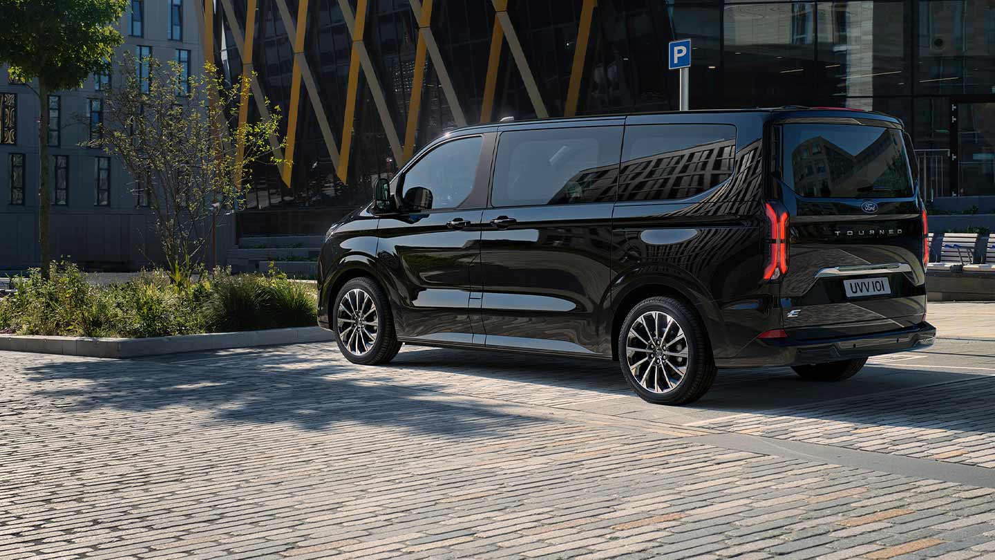 Ford E-Tourneo Custom in Schwarz. Dreiviertelansicht, parkend vor einem Gebäude.